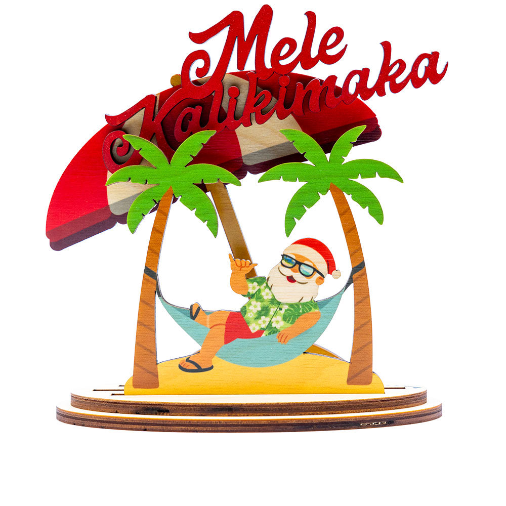 Decorative 3D holiday sign with Santa Claus in a hammock under a red umbrella with palm trees, and 'Mele Kalikimaka' script on a white background.