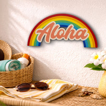 Load image into Gallery viewer, Decorative rainbow sign with 'Aloha' on a wall above a table with beach items.

