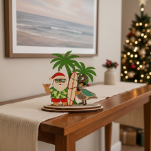 Load image into Gallery viewer, Decorative Santa Claus figurine with a surfboard and nene goose on a wooden table with a Christmas tree in the background.
