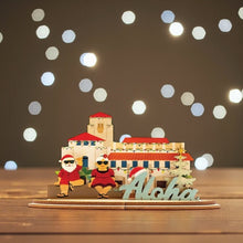 Load image into Gallery viewer, 3D sign of Honolulu Hale building with Shaka Santa and Mrs Clause characters and &#39;Aloha&#39; sign on a wood table with twinkling white lights in background
